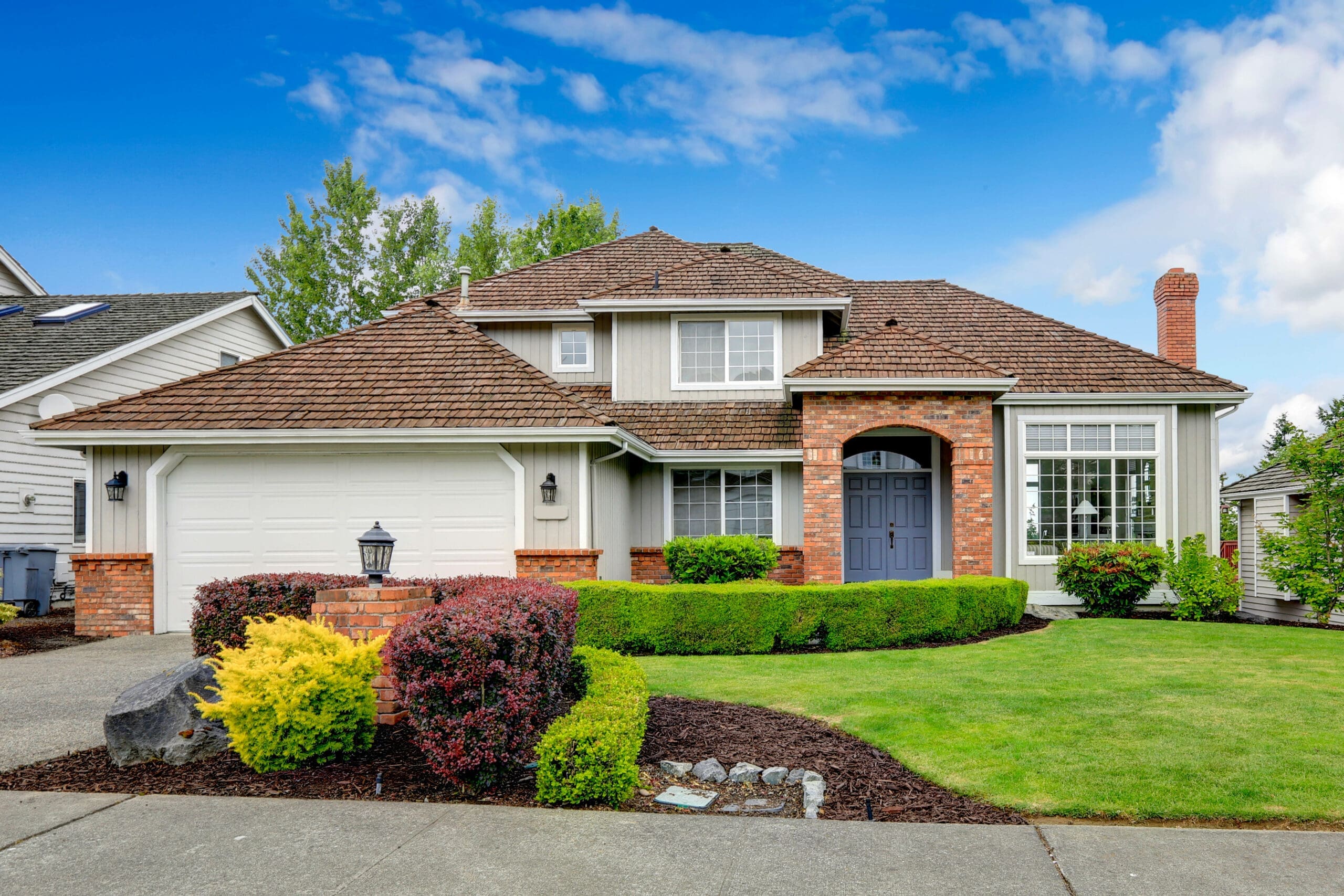 Classic house exterior with brick trimmed entrance porch, green lawn and trimmed hedges. Garage with driveway